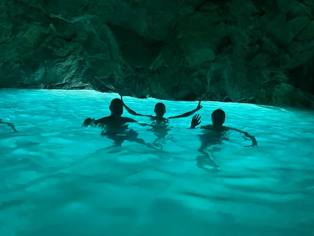 Silhouettes of swimmers inside the Blue Cave in Vlore, standing in crystal-clear turquoise water beneath dramatic limestone formations illuminated by natural light.