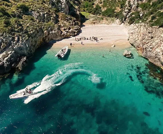 Aerial view of a secluded pebble beach on the Karaburun Peninsula, with crystal-clear turquoise water, visitors relaxing onshore, and speedboats creating white wakes near the coastline surrounded by rocky cliffs.